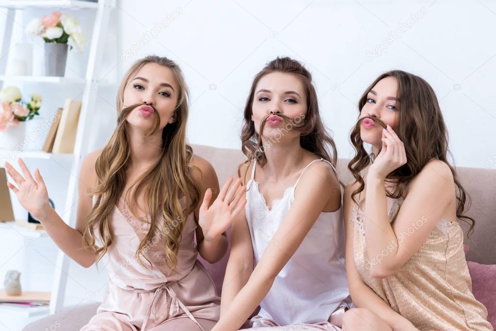 beautiful young women in pajamas holding hair as moustaches and smiling at camera