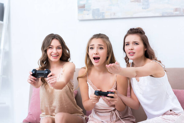 emotional young women in pajamas playing with joysticks together at home
