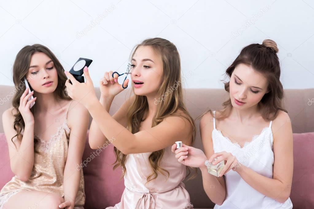 beautiful girlfriends in pajamas applying makeup, holding perfume and talking by smartphone