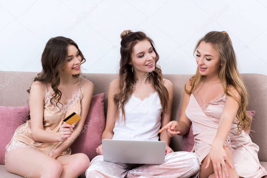 beautiful smiling girlfriends in pajamas using laptop and credit card together