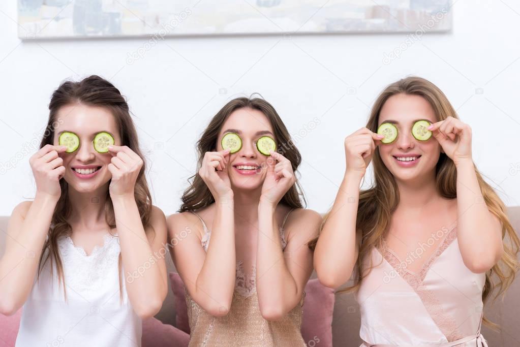 beautiful smiling young women in pajamas holding cucumber slices near eyes