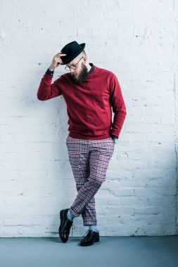 stylish bearded man in eyeglasses with hat standing against white brick wall