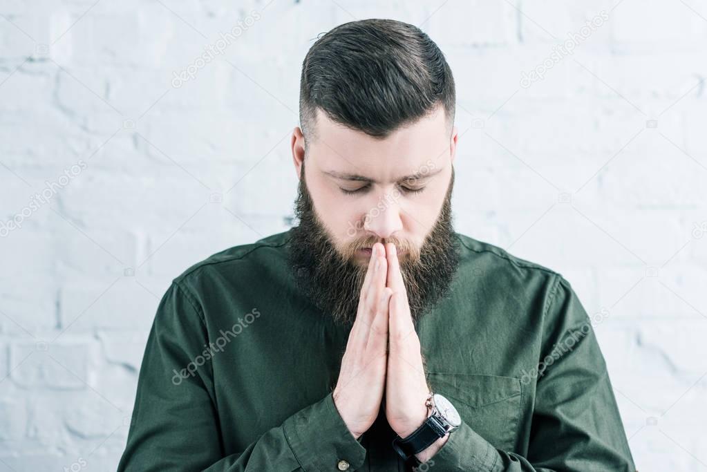 portrait of stylish bearded man praying against white brick wall