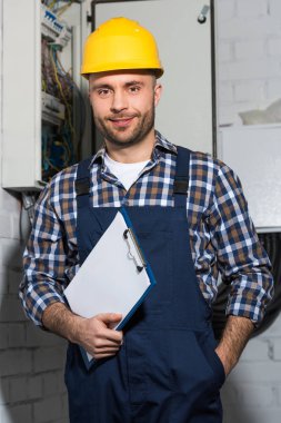 Electrician holding clipboard and smiling by electrical box clipart