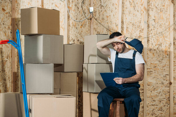 Tired loader man filling checklist while sitting by boxes