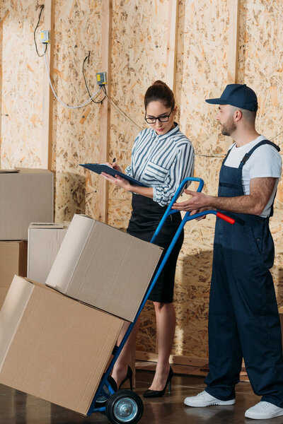 Businesswoman filling checklist while loader man carrying delivery cart