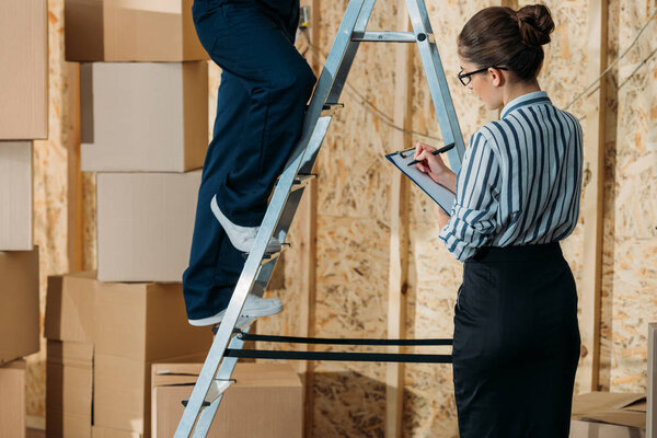 Businesswoman writing in clipboard while loader man standing on a ladder