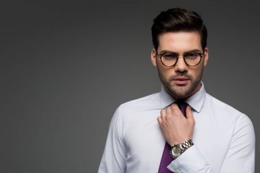 Portrait of businessman in glasses correcting tie isolated on grey clipart