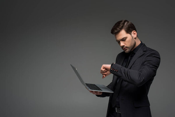 Businessman looking at wristwatch with laptop in hand isolated on grey