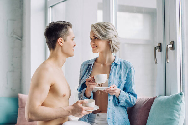 smiling couple looking at each other and holding cups of coffee at home