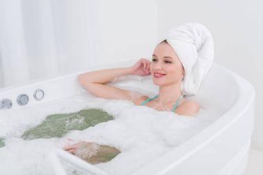smiling woman with towel on head relaxing in bath with foam in spa salon clipart