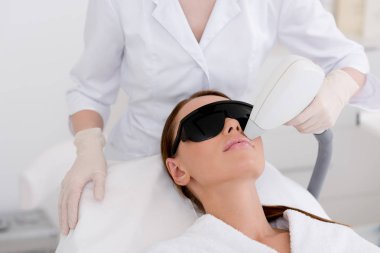 partial view of young woman receiving laser hair removal epilation on face in salon clipart