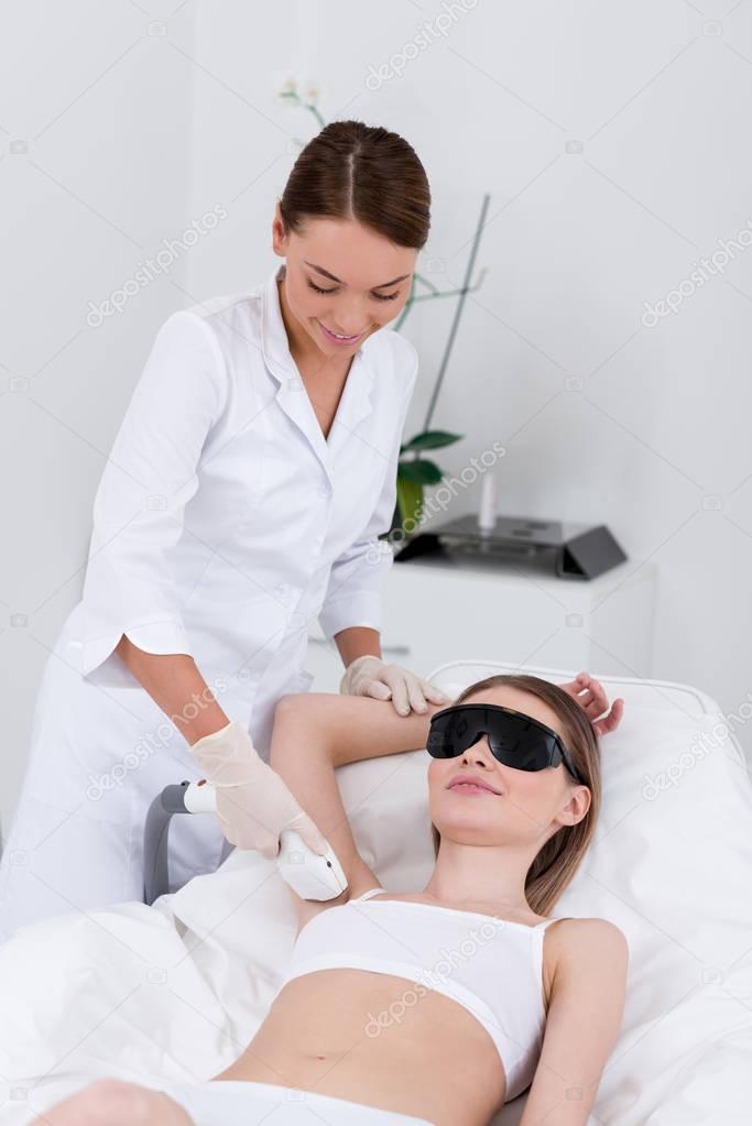 young woman getting laser hair removal epilation on armpit made by cosmetologist in salon