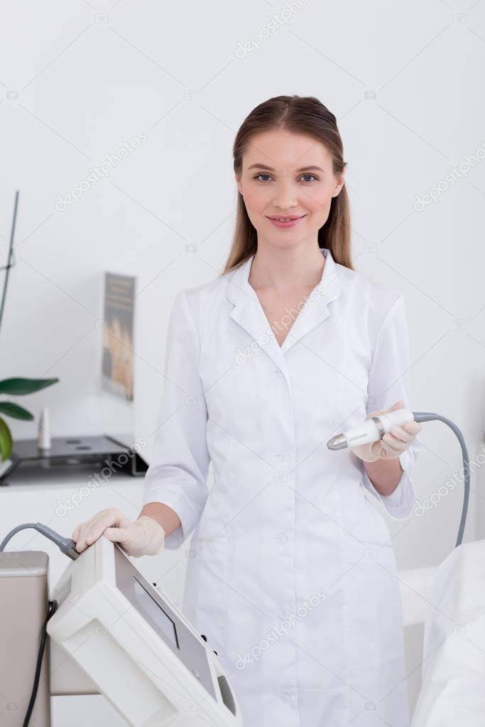 portrait of young cosmetologist in white coat and latex gloves with apparatus in salon