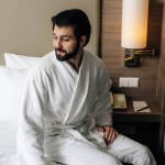 Χαμογελαστός άνθρωπος κάθεται στο κρεβάτι στο suite ξενοδοχείο μπουρνούζι