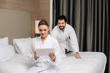 beautiful woman in bathrobe using tablet on bed at hotel suite while her playful boyfriend sneaking to her from behind clipart