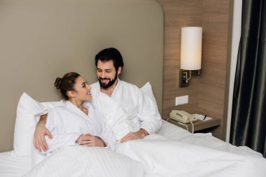 iki otel Süiti yatakta kucaklayan ve birbirine bakarak bornoz içinde gülümseyen