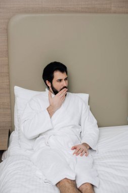 handsome man in bathrobe sitting in bed at hotel suite and yawning clipart