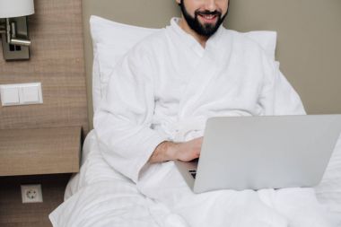 otel odası, yatakta dizüstü kullanarak bornoz erkekte gülümseyen in atış kırpılmış