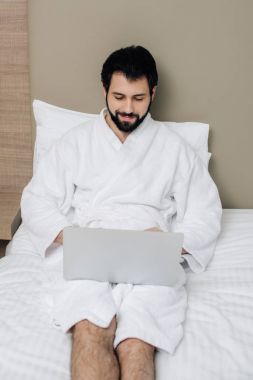 otel odası, yatakta dizüstü kullanarak bornoz gülümseyen adam
