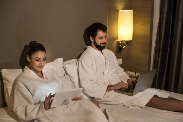 beautiful couple in bathrobes using devices in bed of hotel suite