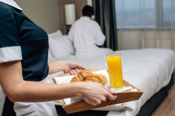 cropped shot of maid in uniform holding tray with croissants and juice for hotel guest