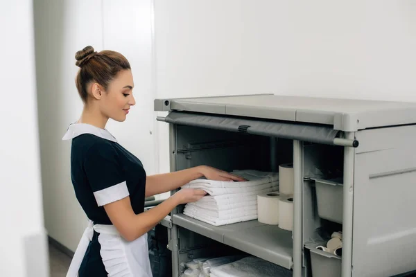 young beautiful maid in uniform taking towels from shelf of housekeeping cart