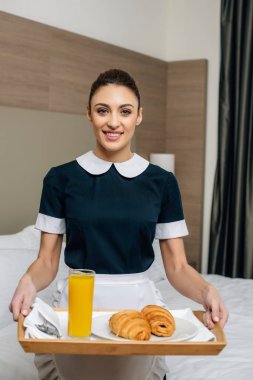 young smiling maid in uniform holding croissants and juice on tray