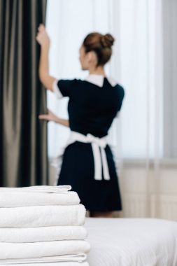 young maid in uniform shutting curtain with stack of clean towels standing on foreground clipart