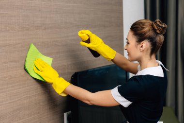 young beautiful maid in uniform wiping wood wall with rag and aerosol furniture cleaner
