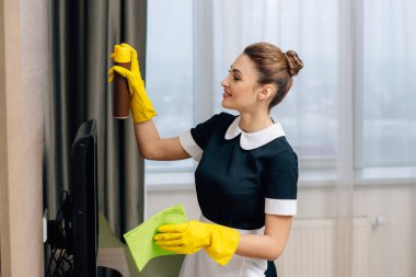genç güzel hizmetçi üniformalı bez ve aerosol mobilya temizleyici temizlik yapıyor