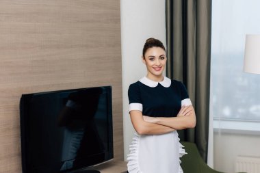 young smiling maid in uniform with crossed arms at hotel suite clipart