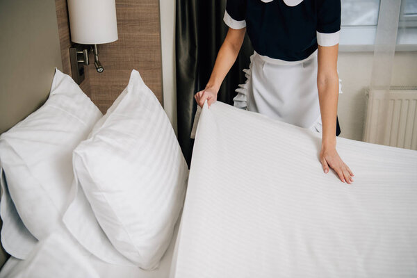 cropped shot of maid in uniform making bed at hotel suite