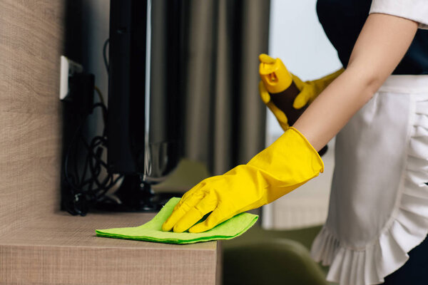 cropped shot of maid in uniform wiping shelf with rag and aerosol furniture cleaner