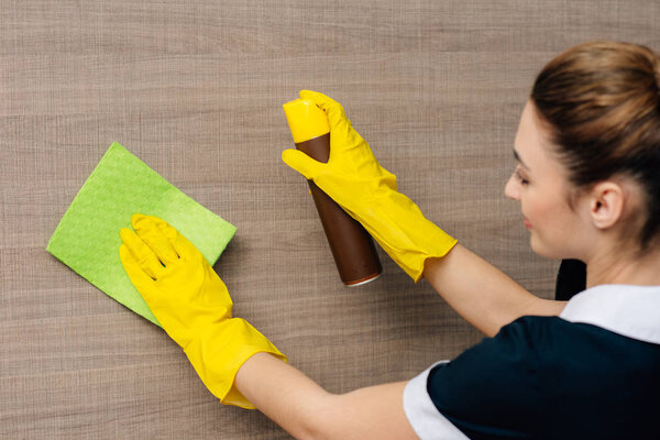 close-up shot of young maid in uniform wiping wood wall with rag and aerosol furniture cleaner