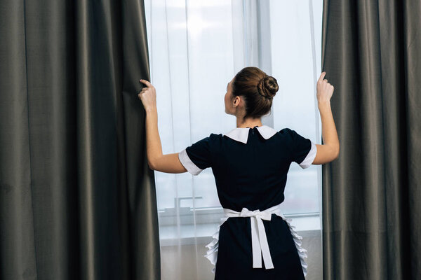 rear view of young beautiful maid in uniform shutting curtains