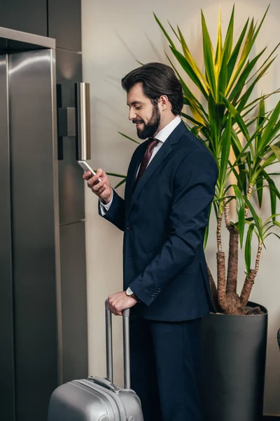 ホテルのエレベーターを待っている間にスマート フォンを使用して荷物を持って笑顔の実業家 — ストック写真