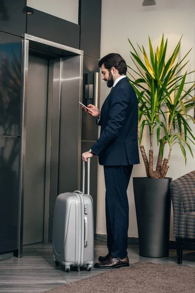 Hombre Negocios Con Equipaje Usando Teléfono Inteligente Mientras Espera Ascensor —  Fotos de Stock