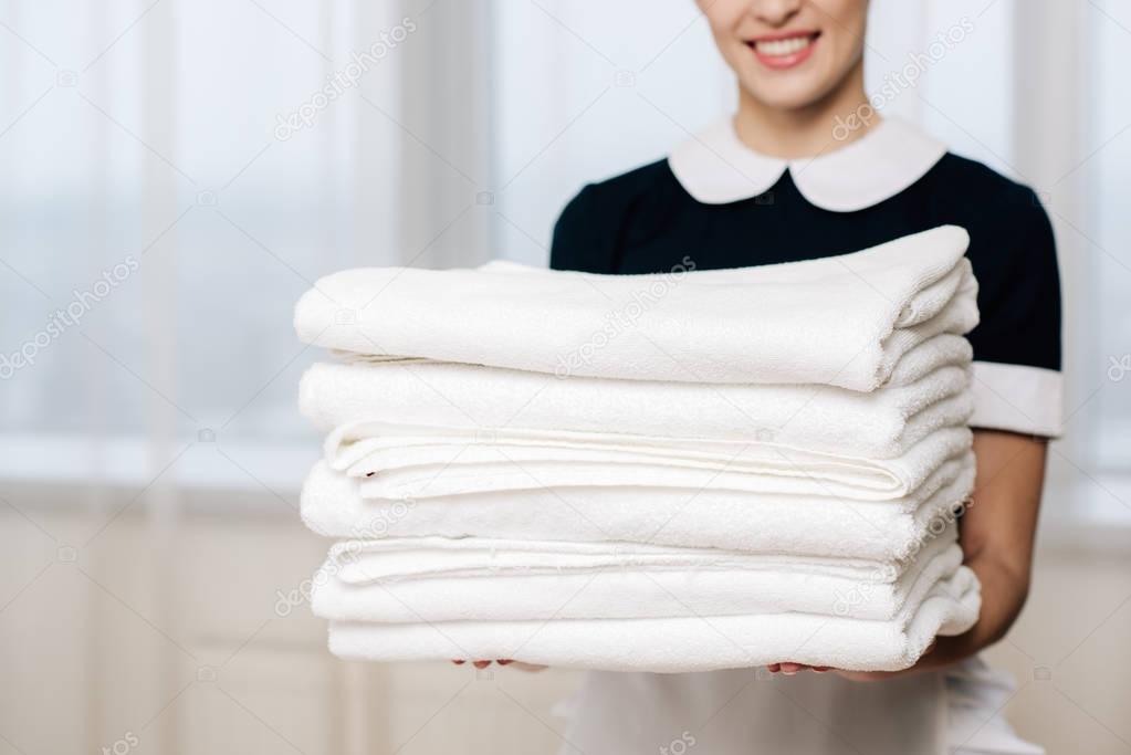 cropped shot of smiling maid in uniform holding stack of clean towels