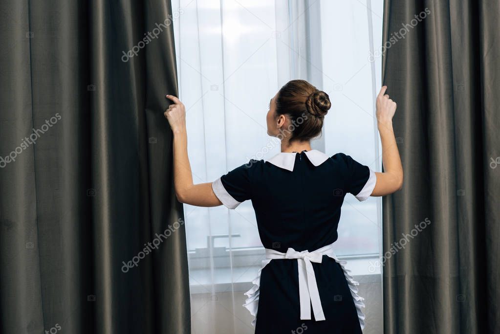 rear view of young beautiful maid in uniform shutting curtains