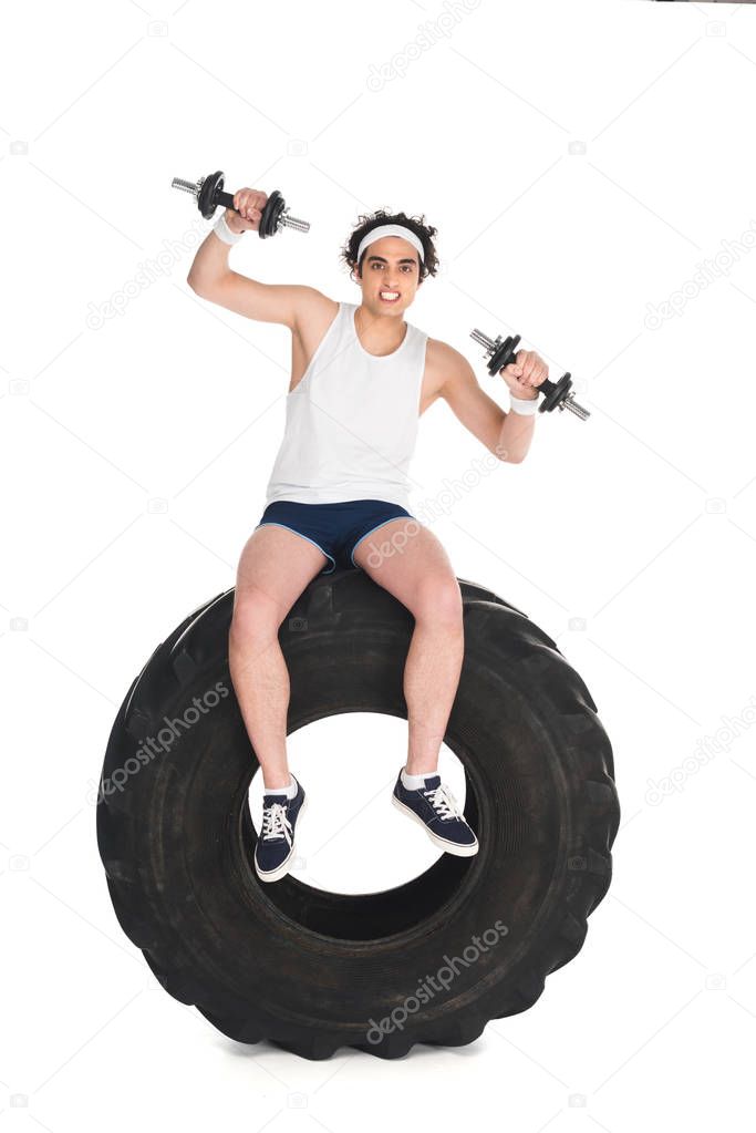 Young thin sportsman with dumbbells sitting on tire of wheel isolated on white