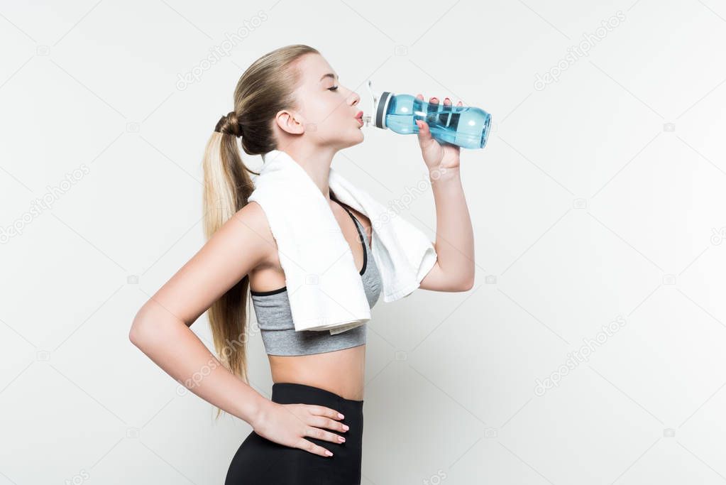 Sportswoman with towel on shoulders drinking from water bottle isolated on white