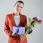 Happy woman holding gift box and flower bouquet isolated on grey