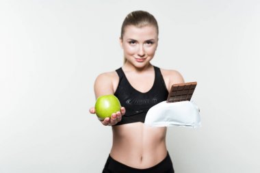 Attractive woman in sports clothes holding apple and chocolate bar isolated on white