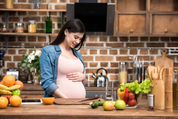 美丽微笑的年轻孕妇抚摸腹部 而在厨房做饭 — 图库照片