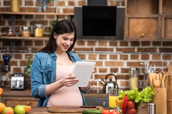 キッチンで料理をしながらデジタル タブレットを使用して笑顔の若い妊婦 — ストック写真