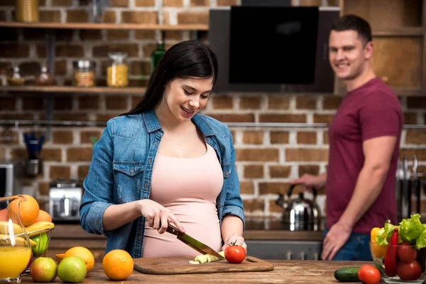 Feliz Jovem Grávida Cortando Legumes Enquanto Sorrindo Marido Atrás Cozinha — Fotografia de Stock