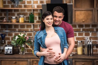 mutlu genç hamile çift kucaklayan ve birlikte evde kameraya gülümseyen