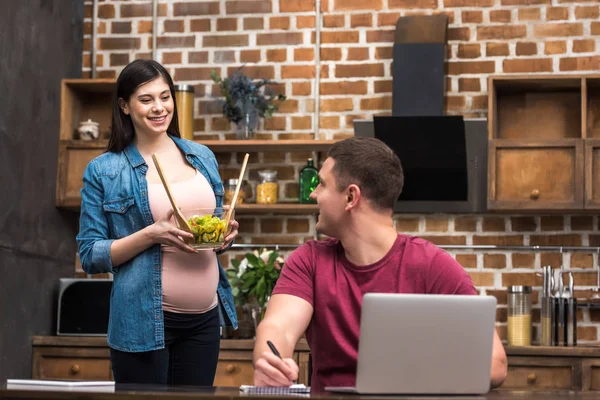 Joven Sonriente Usando Ordenador Portátil Mirando Esposa Embarazada Sosteniendo Tazón — Foto de stock gratuita