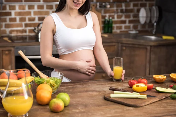 微笑的年轻孕妇抚摸腹部和在厨房里捧着一杯新鲜果汁的镜头 — 图库照片